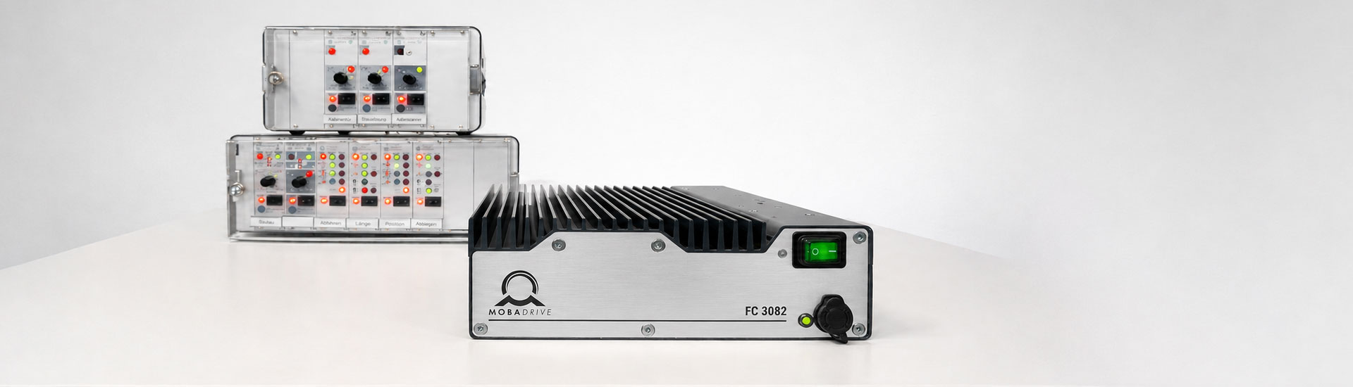 MOBADRIVE FC 3082 frequency controller with brushed metal front panel, green illuminated power switch and black heat sink, shown on a white studio surface with older Schmocker control modules in the background.